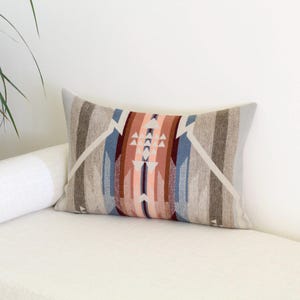 May include: Rectangular cushion with a geometric pattern in shades of grey, brown, blue, and coral. The cushion is placed on a white sofa. The design features vertical stripes and triangular shapes.