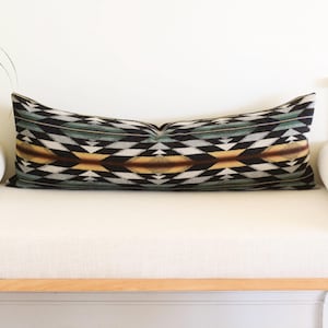 May include: A long, rectangular pillow with a geometric pattern in black, white, teal, and gold. The pillow is placed on a white bench with wooden legs. The pillow's design features diamond and triangle shapes.