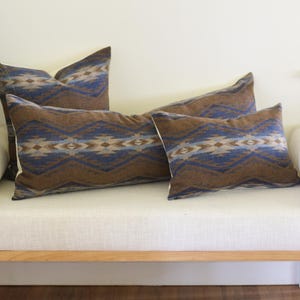 May include: Four decorative cushions with a brown, blue, and cream geometric pattern. The cushions are arranged on a white bench with a light wood frame. The cushions have a soft, textured appearance, suitable for home decor.