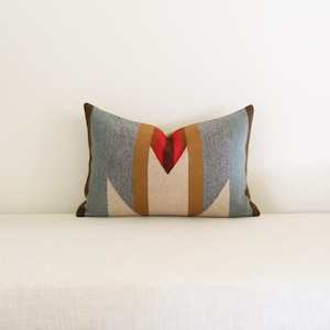 May include: A rectangular throw pillow featuring a geometric pattern in shades of blue, tan, cream, and a central red accent. The pillow has dark brown piping along the edges and a textured fabric finish, centered on a plain white surface.