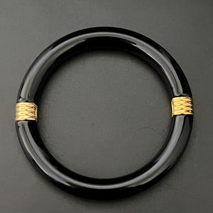 May include: A black bangle bracelet with gold-coloured accents. The bracelet is a solid, glossy black with two gold-coloured bands wrapped around it. The jewellery is set against a dark background.