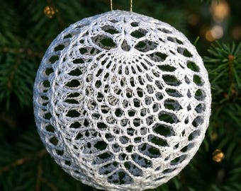 Op de afbeelding: Een bolvormige witte gehaakte decoratie met een ingewikkeld opengewerkt kantpatroon. Het ronde, holle ornament is gemaakt van fijn wit garen en hangt aan een dun gouden koordje, wat een delicate, webachtige textuur laat zien.