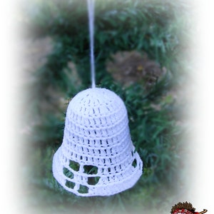 Op de afbeelding: Een witte gehaakte klok als ornament met een opengewerkt patroon langs de onderrand. De klok hangt aan een dun wit draadje en bevat een klein logo in de hoek met de tekst Zicca handmade crochet.