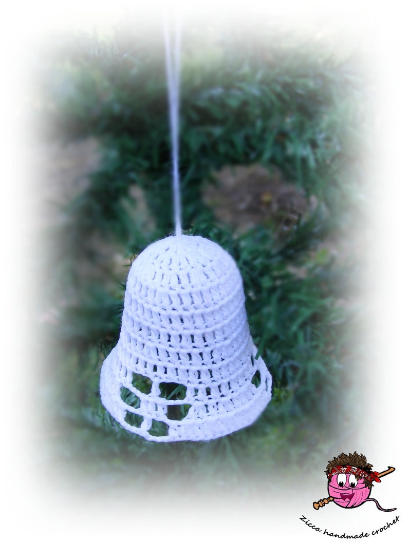 Op de afbeelding: Een witte gehaakte klok als ornament met een opengewerkt patroon langs de onderrand. De klok hangt aan een dun wit draadje en bevat een klein logo in de hoek met de tekst Zicca handmade crochet.
