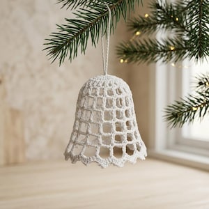 Op de afbeelding: Een wit gehaakt klokornament met een open, rasterachtig patroon en een geschulpte onderrand. De klok hangt aan een dun wit touwtje en toont gedetailleerd haakwerk met een lichtgewicht, holle structuur.