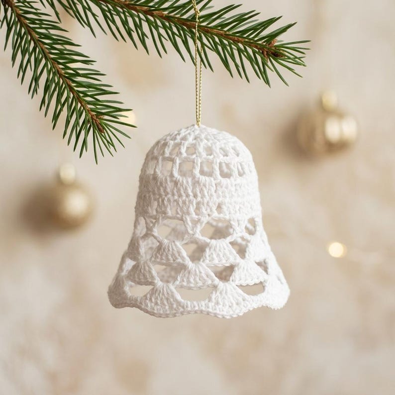 Op de afbeelding: Een witte gehaakte klok als ornament hangend aan een dun gouden koordje. De klok heeft een delicaat opengewerkt patroon met een dicht bovenstuk en een geschulpte onderrand, hangend aan een groene dennentak tegen een wazige achtergrond.
