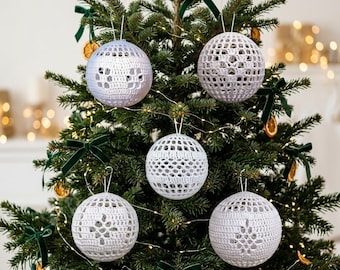 Op de afbeelding: Vijf bolvormige kerstornamenten met witte gehaakte hoezen en ingewikkelde opengewerkte patronen. Elk ornament hangt aan een dun draadje in de takken van een groene dennenboom, versierd met donkergroene fluwelen strikken.