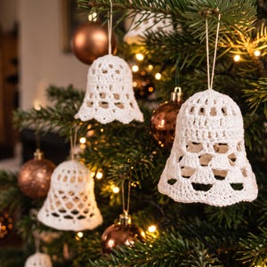 Op de afbeelding: Meerdere witte gehaakte klokornamenten hangen aan de groene takken van een kerstboom. Elke klok heeft een opengewerkt patroon en hangt aan een dun touwtje, afgewisseld met ronde, metallic bronskleurige kerstballen.