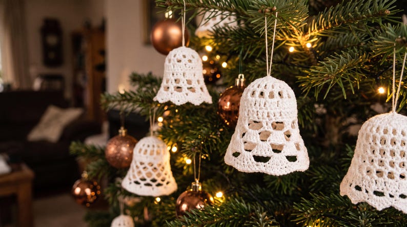 Op de afbeelding: Meerdere witte gehaakte klokornamenten hangen aan de groene takken van een kerstboom. Elke klok heeft een opengewerkt patroon en hangt aan een dun touwtje, afgewisseld met ronde, metallic bronskleurige kerstballen.