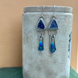 May include: A pair of handmade earrings with a unique design. Each earring features a triangular, blue and white ceramic top, connected to a silver wire and dangling blue beads. The earrings are displayed against a neutral background.
