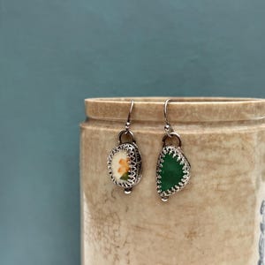 May include: A pair of silver earrings. One earring features a small, oval-shaped design with a floral pattern in orange and yellow. The other earring has a green, semi-circular design. Both earrings have a decorative, scalloped edge.