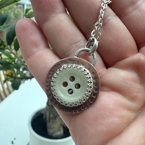 May include: A pendant necklace featuring a central cream-colored four-hole button set within a hammered copper-toned circular frame. The button is bordered by a decorative silver-toned scalloped edge, and the piece hangs from a silver-toned chain.