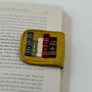 Peut inclure: Un marque-page en feutrine jaune moutarde orné d'une broderie représentant cinq livres verticaux bleu, vert, blanc, rouge et marron foncé. Chaque livre présente des détails cousus blancs sur le dos, le marque-page est plié sur une page.
