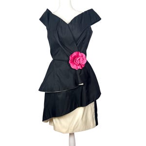 May include: Black off-the-shoulder dress with a layered skirt and a bright pink flower accent at the waist. The dress has a V-neckline and a contrasting cream-colored underskirt. A vintage-inspired formal dress.