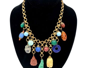 May include: A gold-tone chain necklace featuring a variety of dangling charms, including carved stone beads, spheres, and figures in colors such as green, blue, red, orange, and clear. Each charm is attached to the chain with a small metal cap.