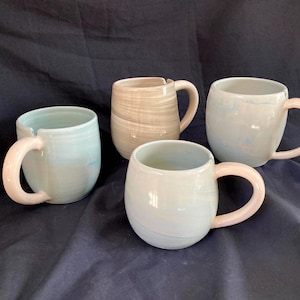 May include: Four ceramic mugs in shades of blue, beige, and white with rounded shapes and large handles. The handles are a light pink color. The mugs are arranged on a dark blue surface.