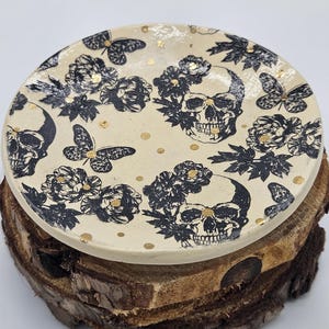 May include: A round, cream-colored ceramic plate featuring a black skull and floral pattern with butterfly accents and gold flecks. The plate is displayed on a rustic, wooden base, creating a contrast of textures and styles. The design is a unique blend of gothic and natural elements.