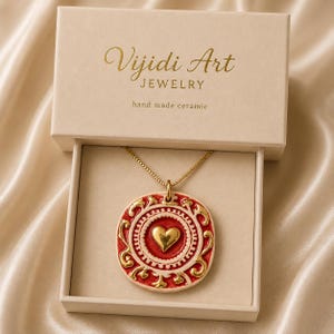 May include: A gold chain necklace with a round ceramic pendant featuring a central gold heart, red and white decorative patterns, and gold accents. The necklace is presented in a beige box labeled Vijidi Art JEWELRY hand made ceramic.