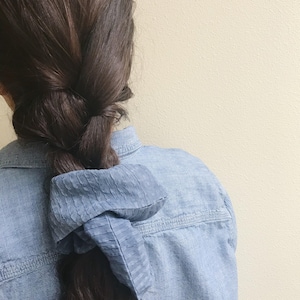 May include: A blue scrunchie securing a dark brown plait. The scrunchie, made of textured fabric, contrasts with the denim jacket. The fabric is a lighter shade of blue.
