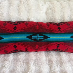May include: A rectangular throw pillow featuring a woven pattern with a prominent central horizontal band in shades of teal and blue. The surrounding areas display a geometric design in deep red and black, incorporating triangular and diamond shapes.