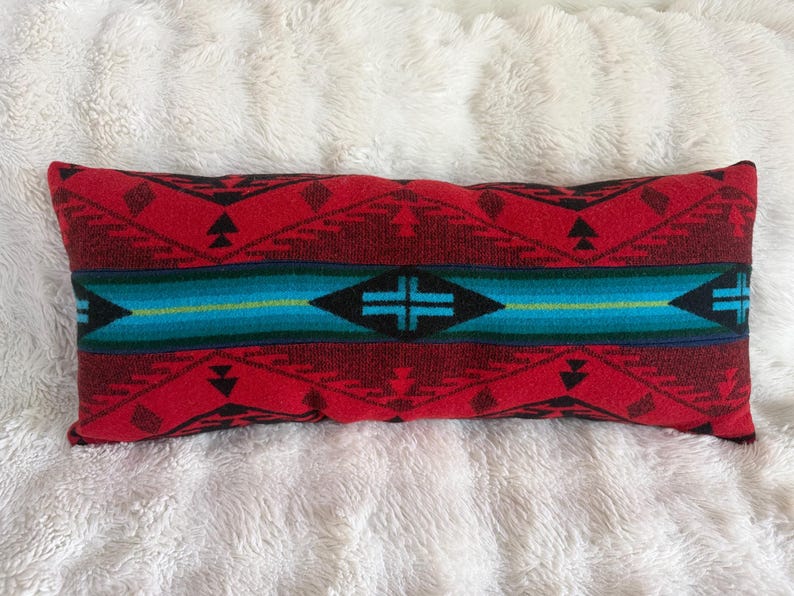 May include: A rectangular throw pillow featuring a woven pattern with a prominent central horizontal band in shades of teal and blue. The surrounding areas display a geometric design in deep red and black, incorporating triangular and diamond shapes.