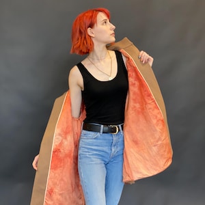 K&ouml;nnte beinhalten: Ein beigefarbener Mantel mit einem leuchtend korallen- und orangefarbenen Tie-Dye-Innenfutter, getragen &uuml;ber einem schwarzen gerippten Tanktop, einem schwarzen Lederg&uuml;rtel mit goldener Schnalle und einer hellblauen Jeans. Der Mantel ist offen.