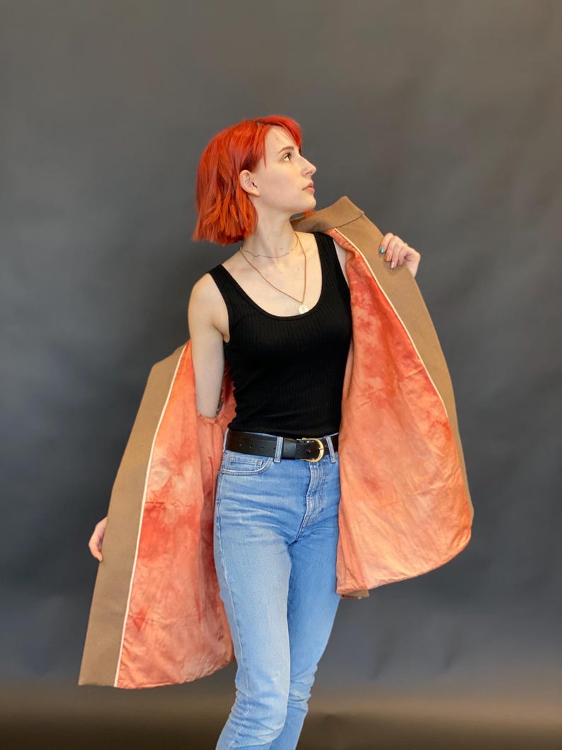 K&ouml;nnte beinhalten: Ein beigefarbener Mantel mit einem leuchtend korallen- und orangefarbenen Tie-Dye-Innenfutter, getragen &uuml;ber einem schwarzen gerippten Tanktop, einem schwarzen Lederg&uuml;rtel mit goldener Schnalle und einer hellblauen Jeans. Der Mantel ist offen.