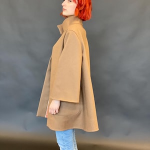 K&ouml;nnte beinhalten: Ein beigefarbener, mittellanger Mantel mit hohem Kragen, weiten Dreiviertel&auml;rmeln und zwei gro&szlig;en aufgesetzten Taschen auf der Vorderseite. Der Mantel hat eine ausgestellte A-Linien-Silhouette und wird &uuml;ber einer hellblauen Jeans getragen.