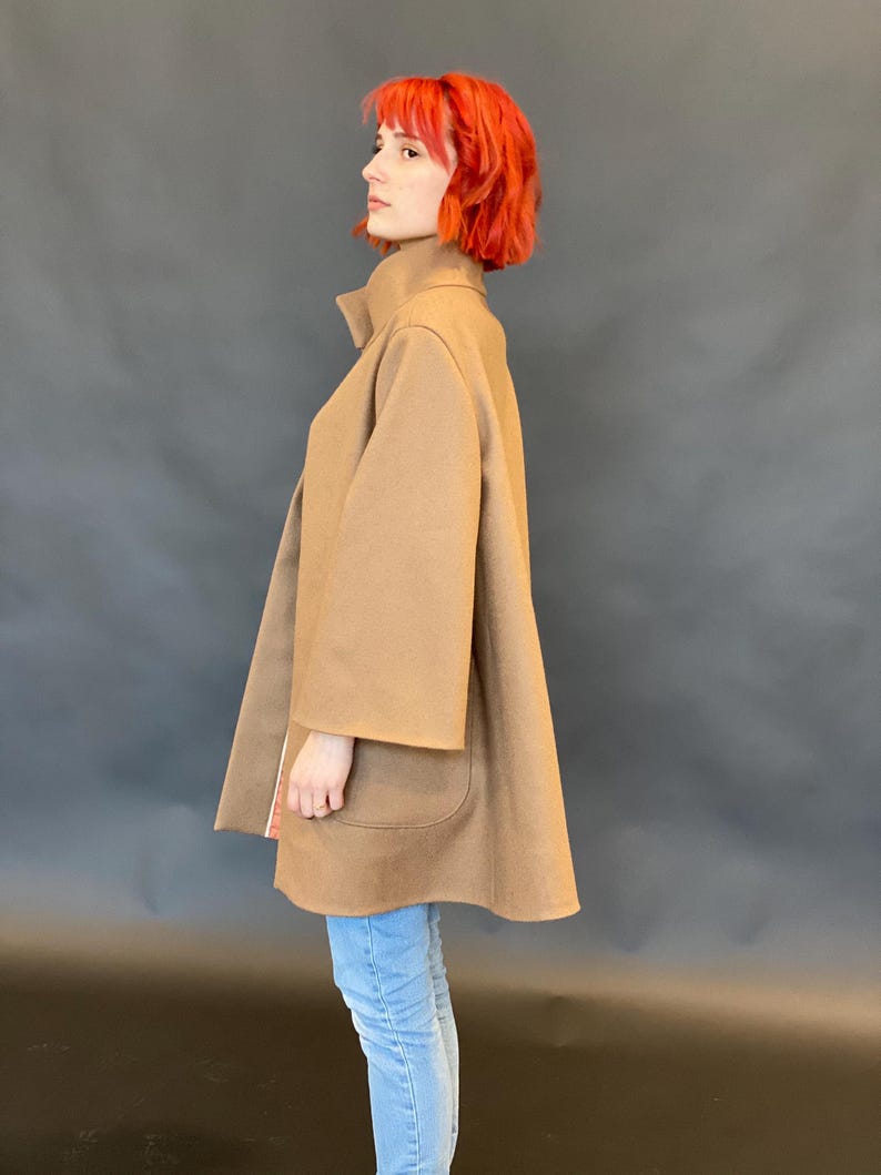 K&ouml;nnte beinhalten: Ein beigefarbener, mittellanger Mantel mit hohem Kragen, weiten Dreiviertel&auml;rmeln und zwei gro&szlig;en aufgesetzten Taschen auf der Vorderseite. Der Mantel hat eine ausgestellte A-Linien-Silhouette und wird &uuml;ber einer hellblauen Jeans getragen.