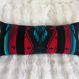 May include: A rectangular lumbar pillow featuring a bold geometric pattern in black, vibrant red, and bright turquoise. The design consists of symmetrical triangular and chevron motifs arranged in vertical bands across the length of the cushion.