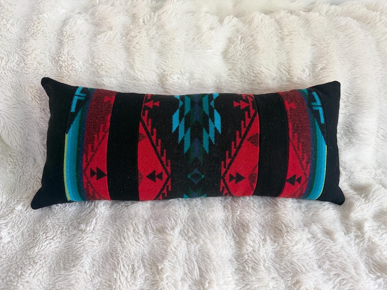 May include: A rectangular lumbar pillow featuring a bold geometric pattern in black, vibrant red, and bright turquoise. The design consists of symmetrical triangular and chevron motifs arranged in vertical bands across the length of the cushion.
