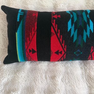May include: A rectangular throw pillow featuring a black base with vertical panels of red, turquoise, and dark blue geometric patterns. The design includes repeating triangular shapes and linear motifs across the fabric surface.
