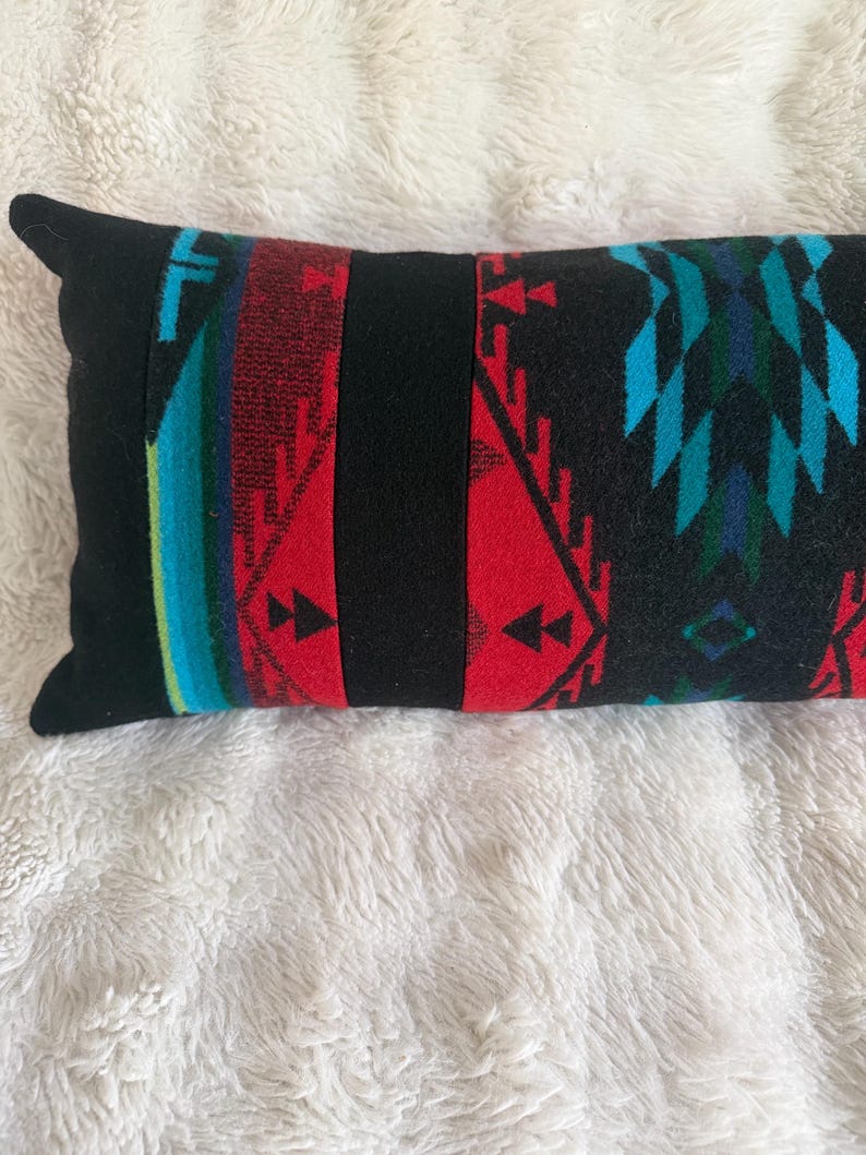 May include: A rectangular throw pillow featuring a black base with vertical panels of red, turquoise, and dark blue geometric patterns. The design includes repeating triangular shapes and linear motifs across the fabric surface.