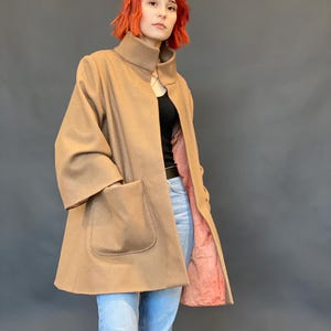 Op de afbeelding: Een beige jas met een hoge kraag, wijde driekwartmouwen en twee grote opgestikte zakken aan de voorzijde. De jas heeft een zichtbare roze voering aan de binnenzijde, een losse pasvorm en een gladde textuur, gedragen over een zwart topje en een blauwe spijkerbroek.