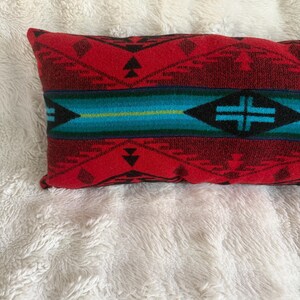 May include: A rectangular throw pillow featuring a woven pattern in shades of red, black, and turquoise. The design includes geometric shapes, triangles, and a central horizontal band with a cross motif, all rendered in a textured fabric weave.
