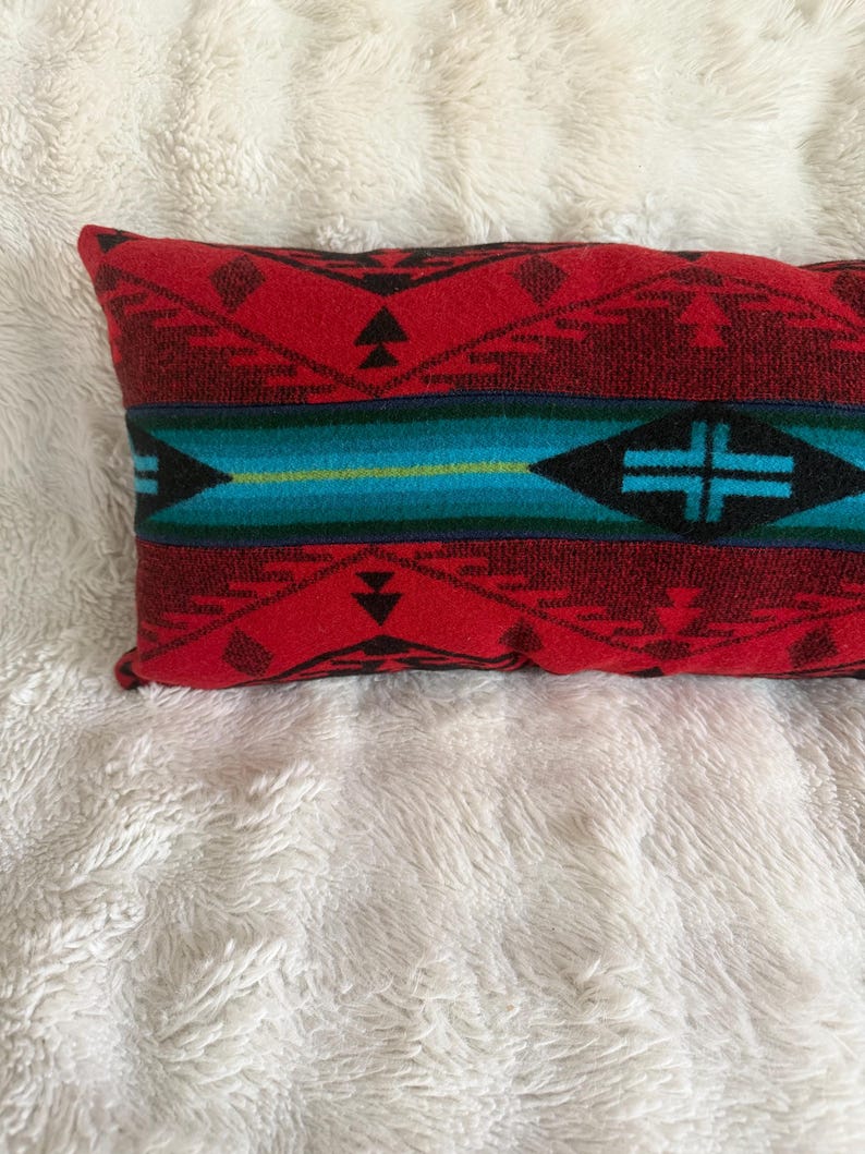 May include: A rectangular throw pillow featuring a woven pattern in shades of red, black, and turquoise. The design includes geometric shapes, triangles, and a central horizontal band with a cross motif, all rendered in a textured fabric weave.