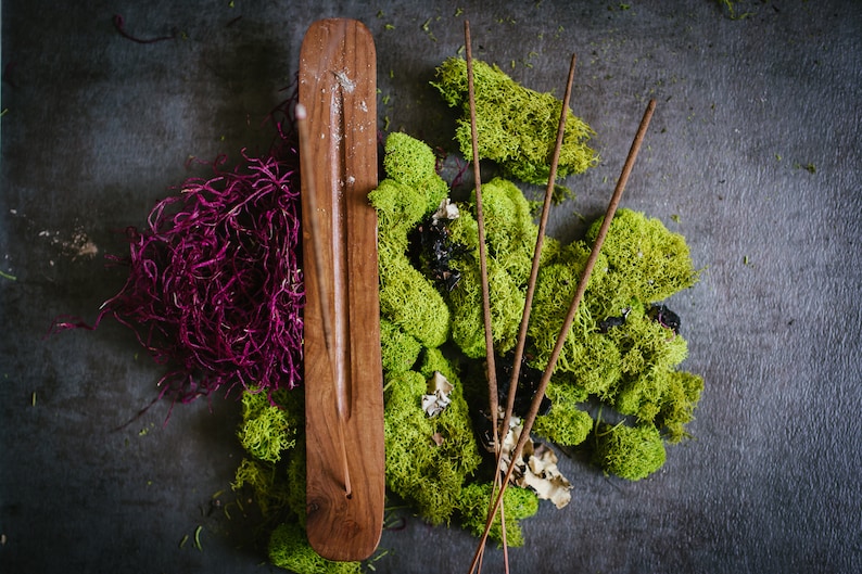 Puede incluir: Un porta incienso de madera con detalles plateados, rodeado de musgo verde, algas moradas y varillas de incienso. La disposici&oacute;n se encuentra sobre una superficie texturizada gris oscuro, creando una est&eacute;tica natural y terrosa.
