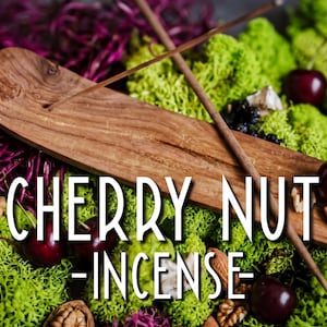 May include: A wooden incense holder with a long, thin incense stick resting on top. The holder is surrounded by bright green moss, dark red cherries, and brown walnuts. The text CHERRY NUT -INCENSE- is overlaid in white capital letters.