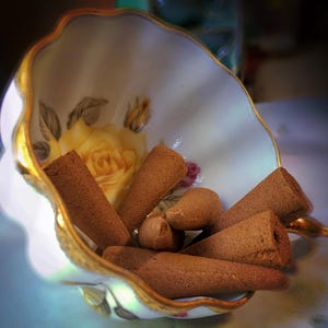 Pode incluir: Uma ch&aacute;vena de ch&aacute; branca com guarni&ccedil;&atilde;o dourada e desenhos florais cont&eacute;m uma cole&ccedil;&atilde;o de cones de incenso castanhos.