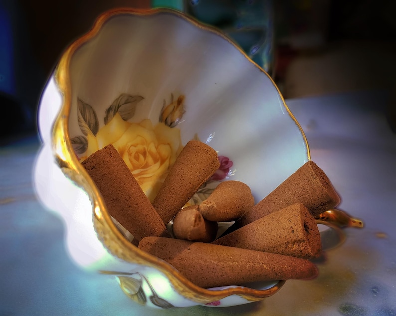 Pode incluir: Uma ch&aacute;vena de ch&aacute; branca com guarni&ccedil;&atilde;o dourada e desenhos florais cont&eacute;m uma cole&ccedil;&atilde;o de cones de incenso castanhos.