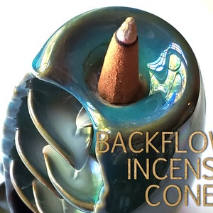 Puede incluir: Un porta conos de incienso de reflujo de color verde azulado y dorado con forma de concha de nautilus. Un cono de incienso marr&oacute;n est&aacute; colocado en el soporte. Las palabras "BACKFLOW INCENSE CONES" est&aacute;n impresas en dorado.