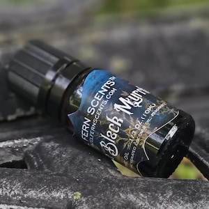 Può includere: Un piccolo flacone in vetro ambrato scuro con tappo nero e un'etichetta con la scritta Fern Scents Black Myrrh, 1/3 oz (10 ml), olio di cocco frazionato e fragranza. Il flacone è posizionato orizzontalmente, mostrando il liquido scuro all'interno.