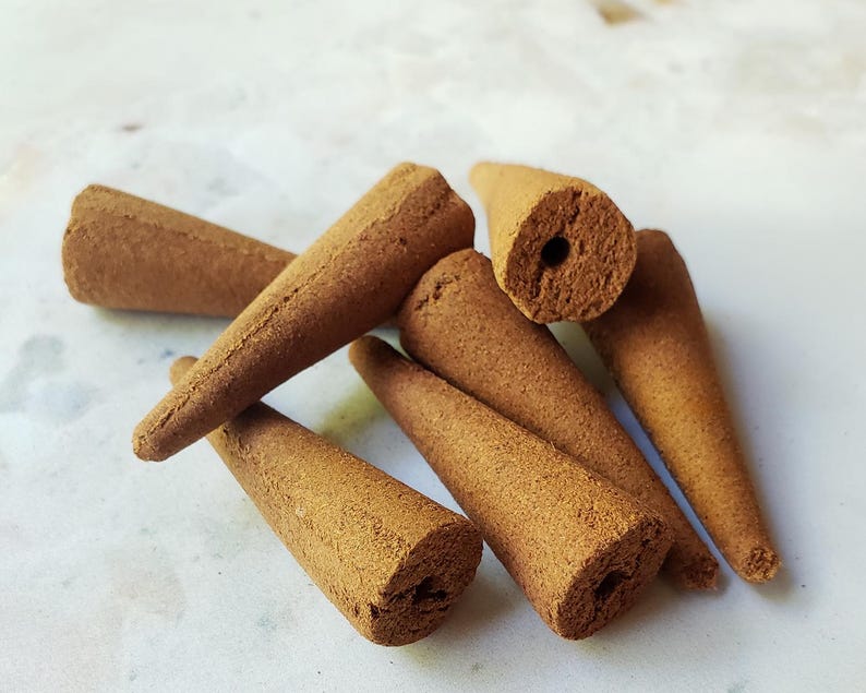 May include: A group of six brown incense cones, each with a small hole in the center. The cones are arranged in a pile on a white surface.