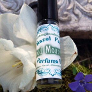 May include: A small glass bottle with a black cap and a white label. The label has green text that reads "Hazel Feather Wood Mountain Perfume Oil".