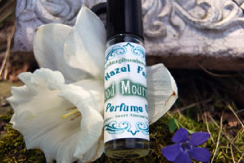 May include: A small glass bottle with a black cap and a white label. The label has green text that reads "Hazel Feather Wood Mountain Perfume Oil".