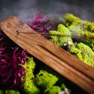 Puede incluir: Un porta incienso de madera de color marr&oacute;n oscuro. Se encuentra sobre un lecho de musgo verde vibrante y follaje p&uacute;rpura intenso. Un palito de incienso est&aacute; encendido, emitiendo una delicada estela de humo. La imagen tiene una est&eacute;tica natural y terrosa.