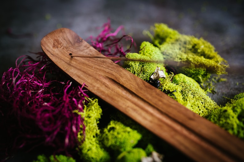Puede incluir: Un porta incienso de madera de color marr&oacute;n oscuro. Se encuentra sobre un lecho de musgo verde vibrante y follaje p&uacute;rpura intenso. Un palito de incienso est&aacute; encendido, emitiendo una delicada estela de humo. La imagen tiene una est&eacute;tica natural y terrosa.