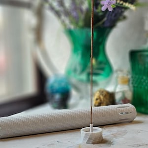 May include: White ceramic incense holder with a long, thin incense stick. The holder has a cylindrical shape with a textured surface and a small hole in the center. The incense stick is positioned vertically in the holder.