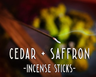 Può includere: Primo piano di un bastoncino di incenso fumante con una punta bianca, che emette una nuvola di fumo. Il testo "CEDAR + SAFFRON - INCENSE STICKS -" è visibile in primo piano.