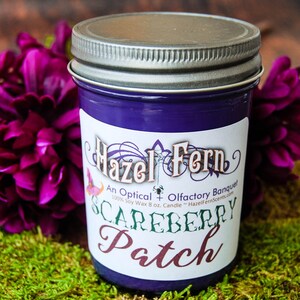 May include: A purple candle in a glass jar with a silver lid. The label reads "Hazel Fern Scareberry Patch". The candle is surrounded by purple flowers and green moss.